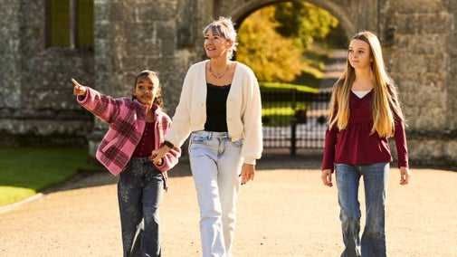 A woman and two girls walk together in front of a stone archway at Lanhydrock. The younger girl points excitedly towards something out of view while holding the woman's hand.